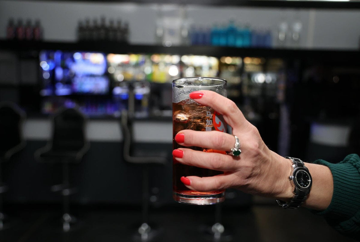 Une main tenant un verre devant le comptoir éclairé d’un bar.